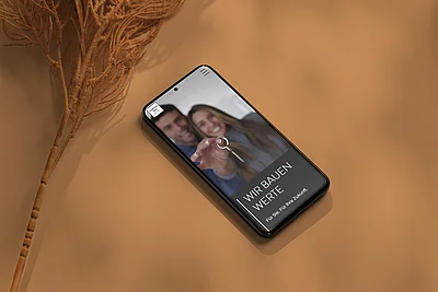 Smartphone auf beigem Untergrund mit Immobilien-App, auf dem Bildschirm ein Paar und der Text „Wir bauen Werte“
