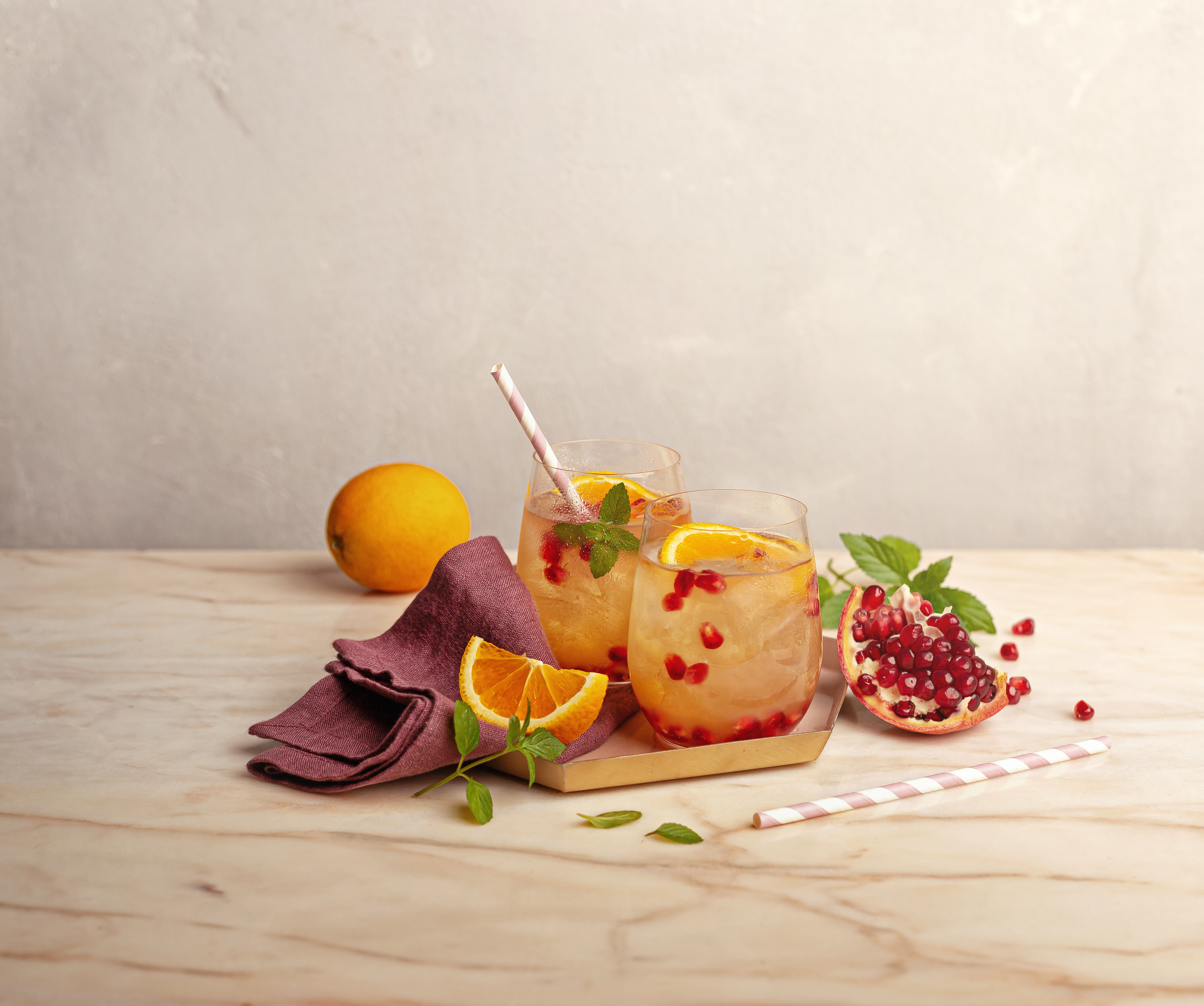 Zwei Gläser mit erfrischendem Orangen-Granatapfel-Getränk, garniert mit Minze, Eiswürfeln und Strohhalmen auf hellem Tisch
