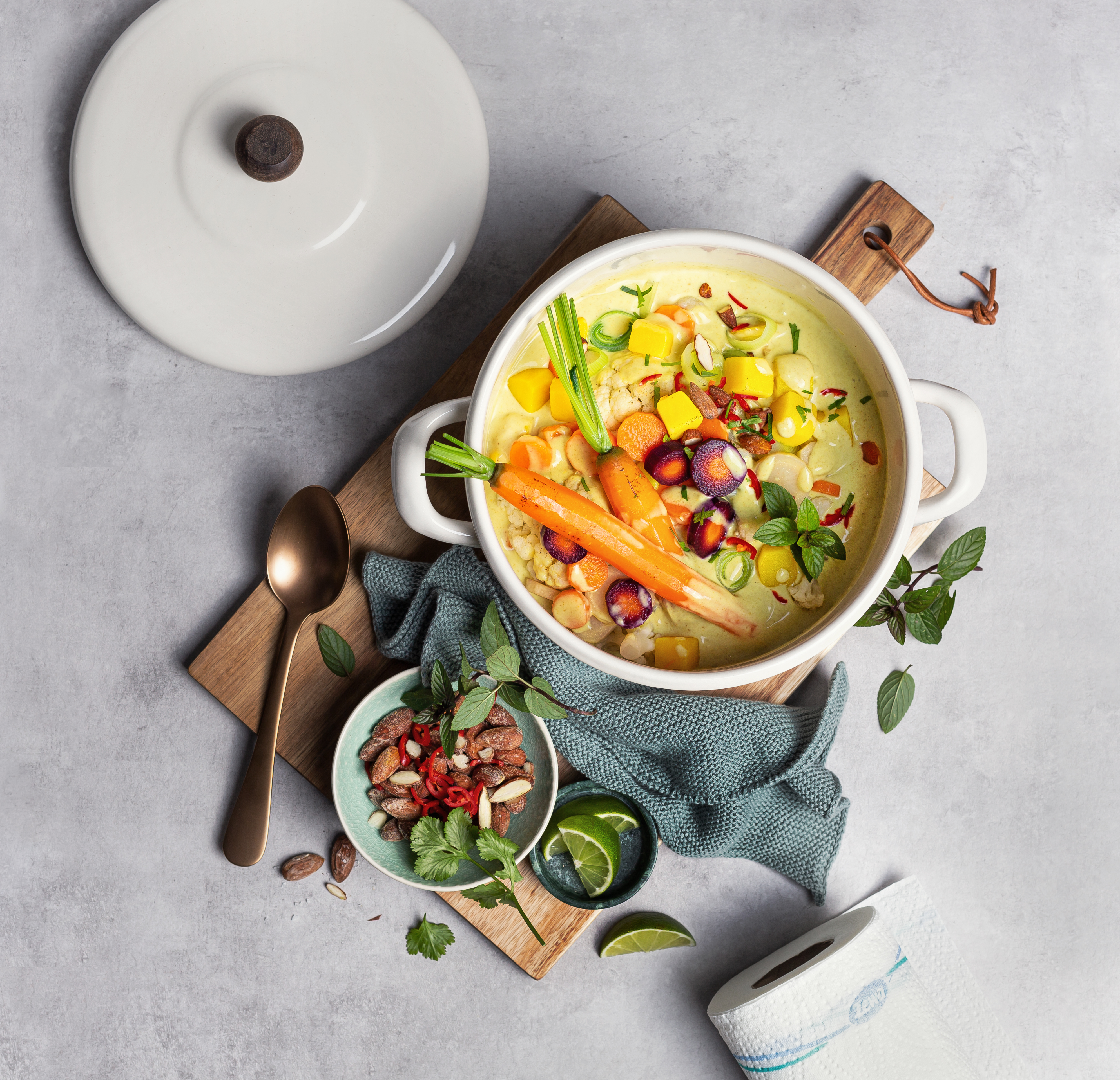 Topf mit bunter Gemüsesuppe aus Karotten, Kartoffeln und Hülsenfrüchten auf grauer Oberfläche, umgeben von Kräutern und Zutaten