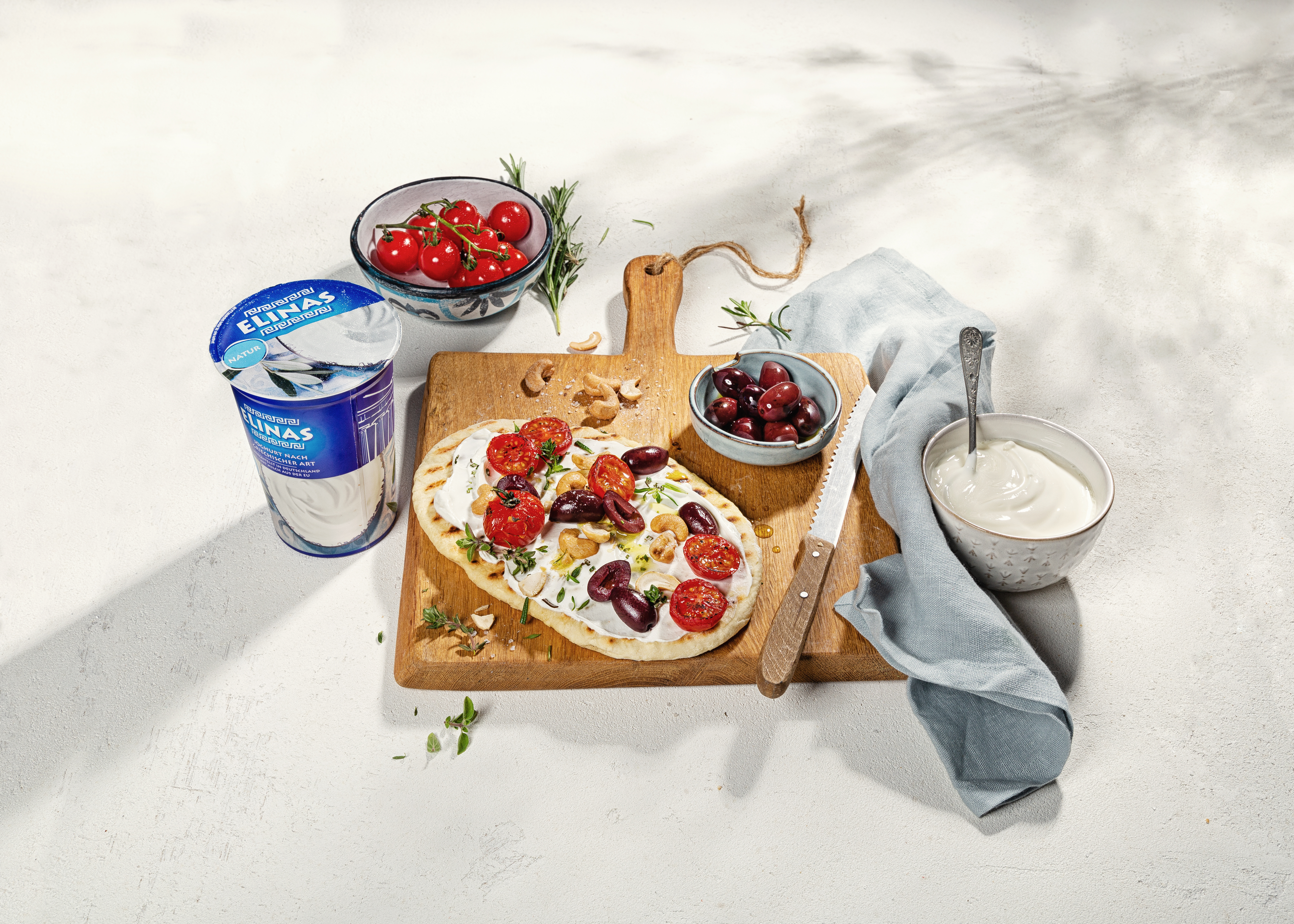 Fladenbrot mit Joghurt, Frischkäse, Kirschtomaten, Oliven und Nüssen auf Holzbrett, umgeben von Schalen mit Zutaten