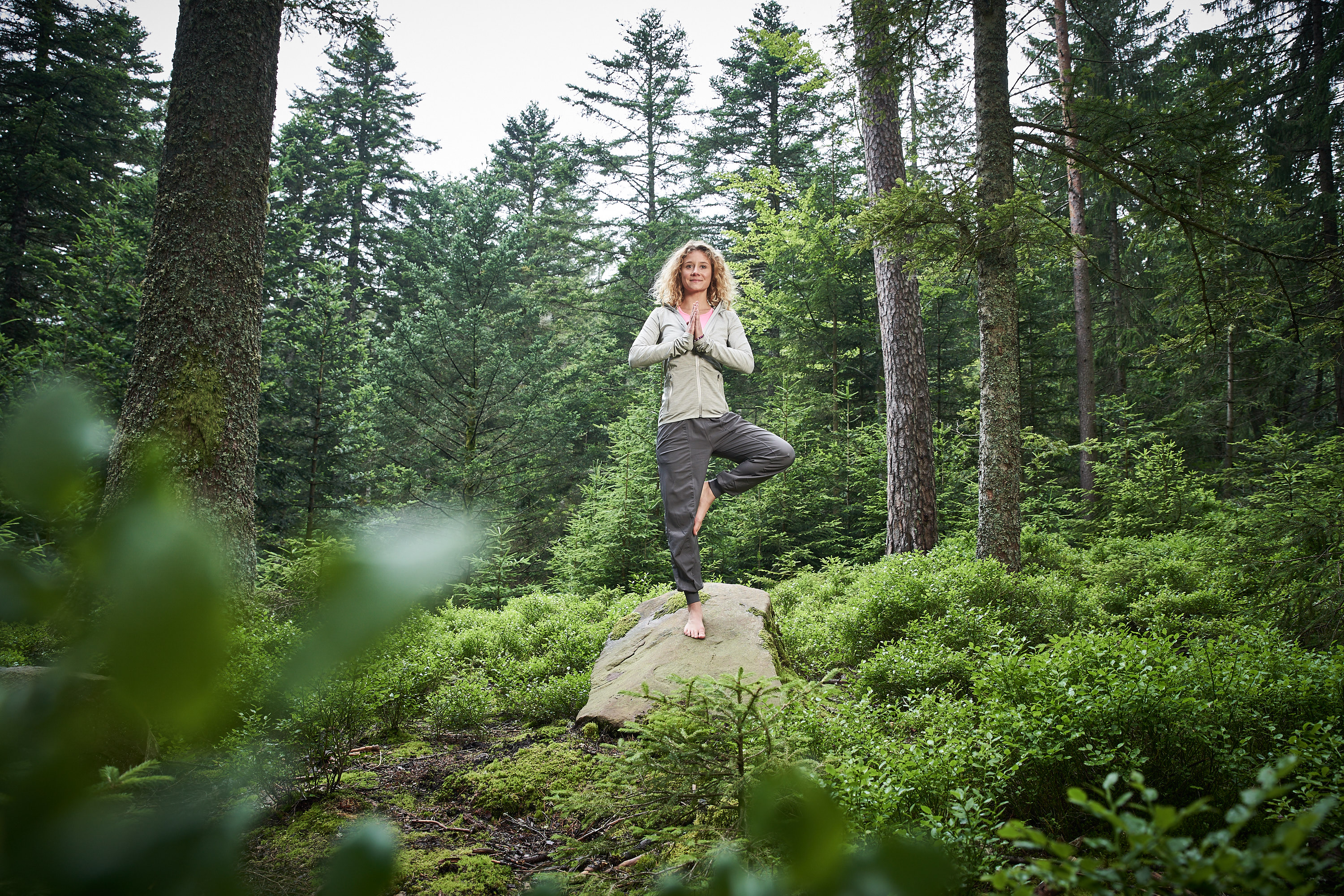 Person steht barfuß im Yoga-Baumstand auf einem Felsen in einem dichten, grünen Wald und hält die Hände vor der Brust zusammen.