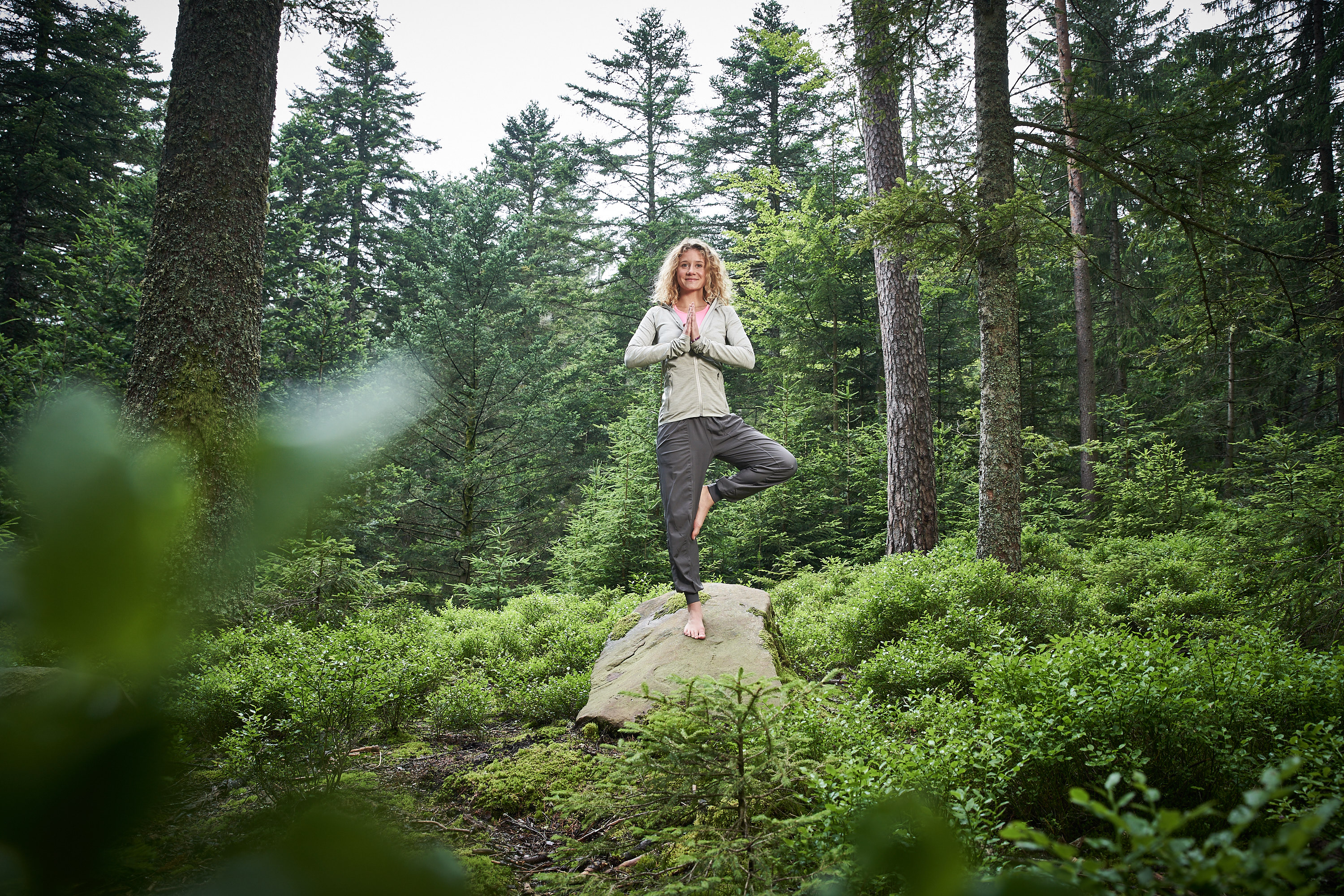 Person steht barfuß im Yoga-Baumstand auf einem Felsen in einem dichten, grünen Wald und hält die Hände vor der Brust zusammen.