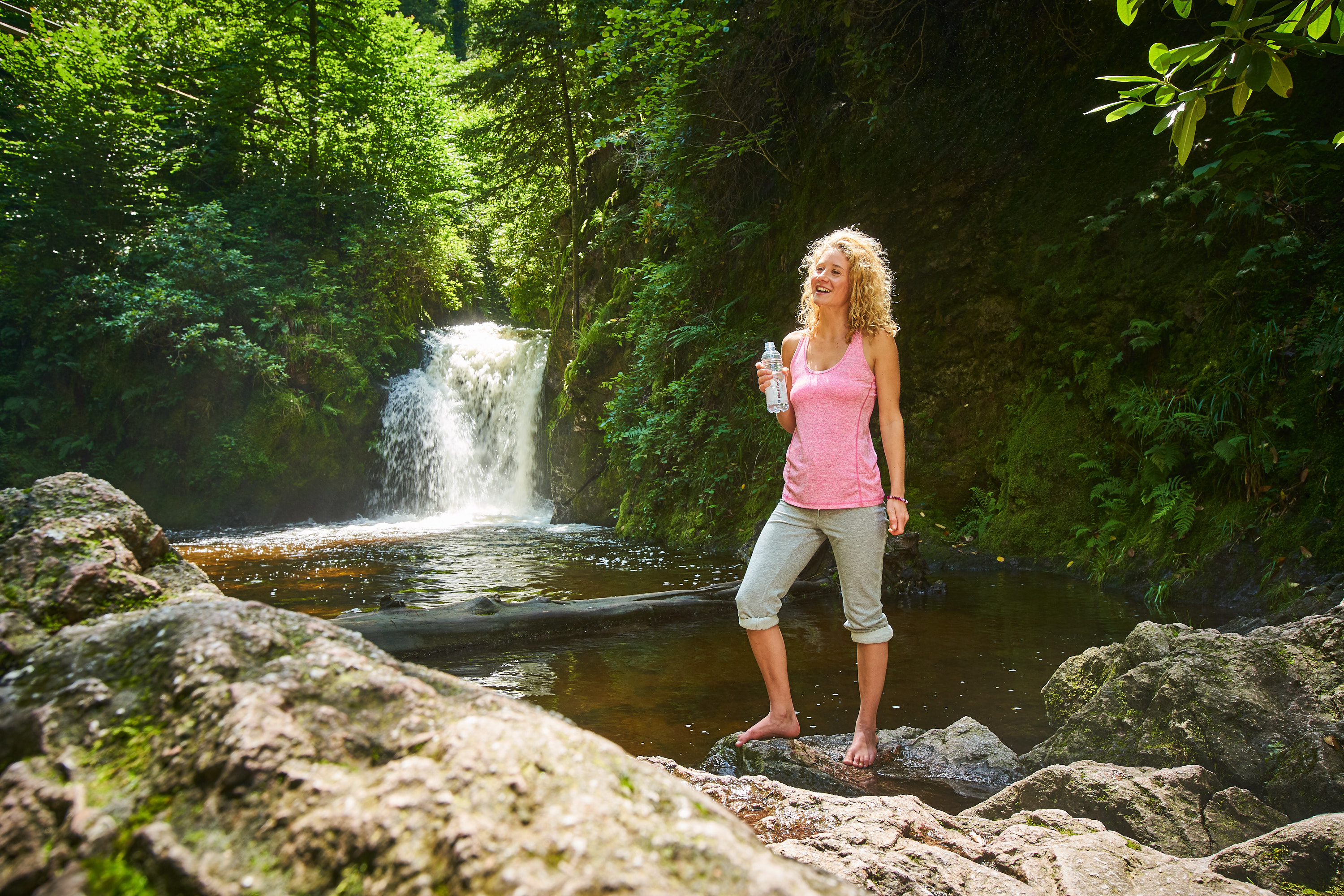 Junge Frau in sportlicher Kleidung hält eine Flasche Black Forest still und steht mit beiden Füßen in einem Gewässer vor einem kleinen Wasserfall im Wald