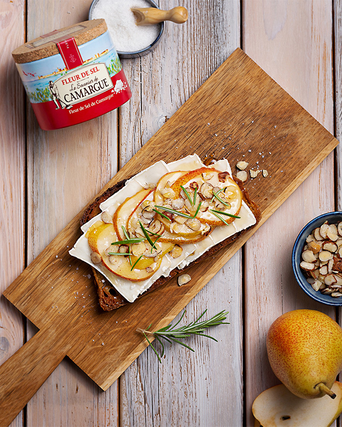 Belegtes Brot mit Birnenscheiben, Käse und Nüssen auf Holzbrett, daneben eine Packung Fleur de Sel von Le Saunier de Camargue von oben