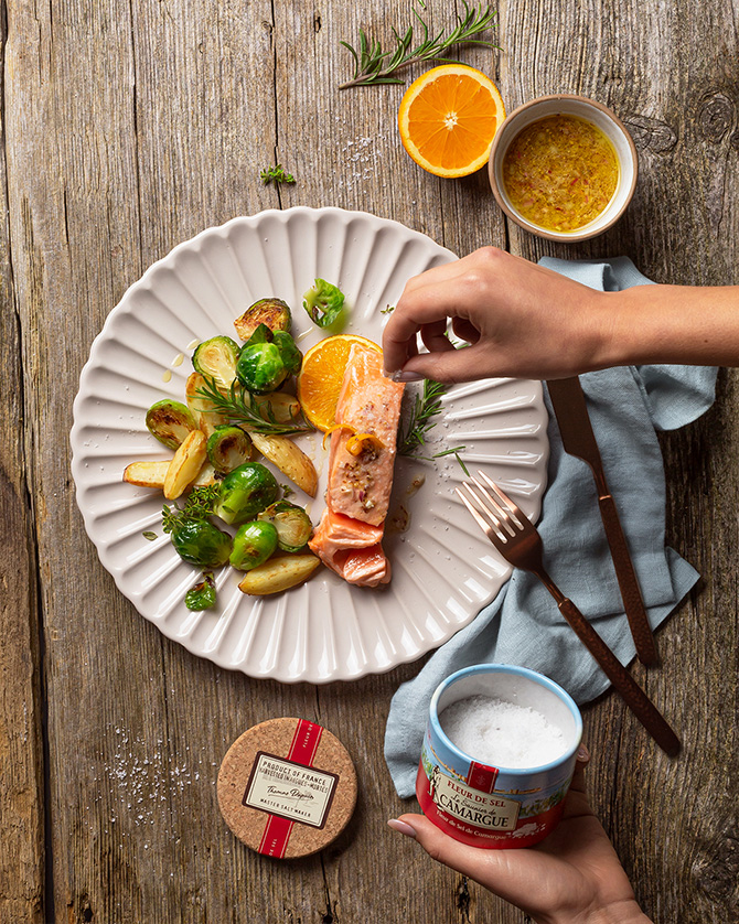 Gebratener Lachs mit Rosenkohl und Orangenfilets auf weißem Teller wird mit Fleur de Sel von Le Saunier de Camargue bestreut in der Draufsicht