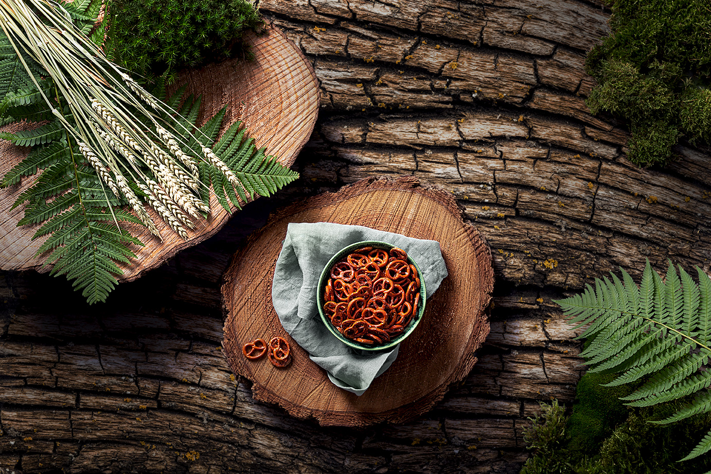 Foto von der Draufsicht auf eine Schale MAYKA Bio-Mini-Brezeln auf einer Holzscheibe. Daneben liegen Strohalme und Farnblätter. Der Untergrund ist eine grobe Rindenstruktur.