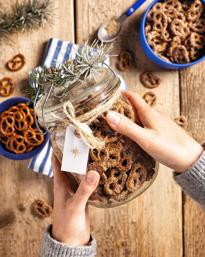 Zwei Hände halten ein großes Einmachglas gefüllt mit gebrannten Brezlen. An dem Glas hängt ein Geschenkeetikett an einer groben Kordel.