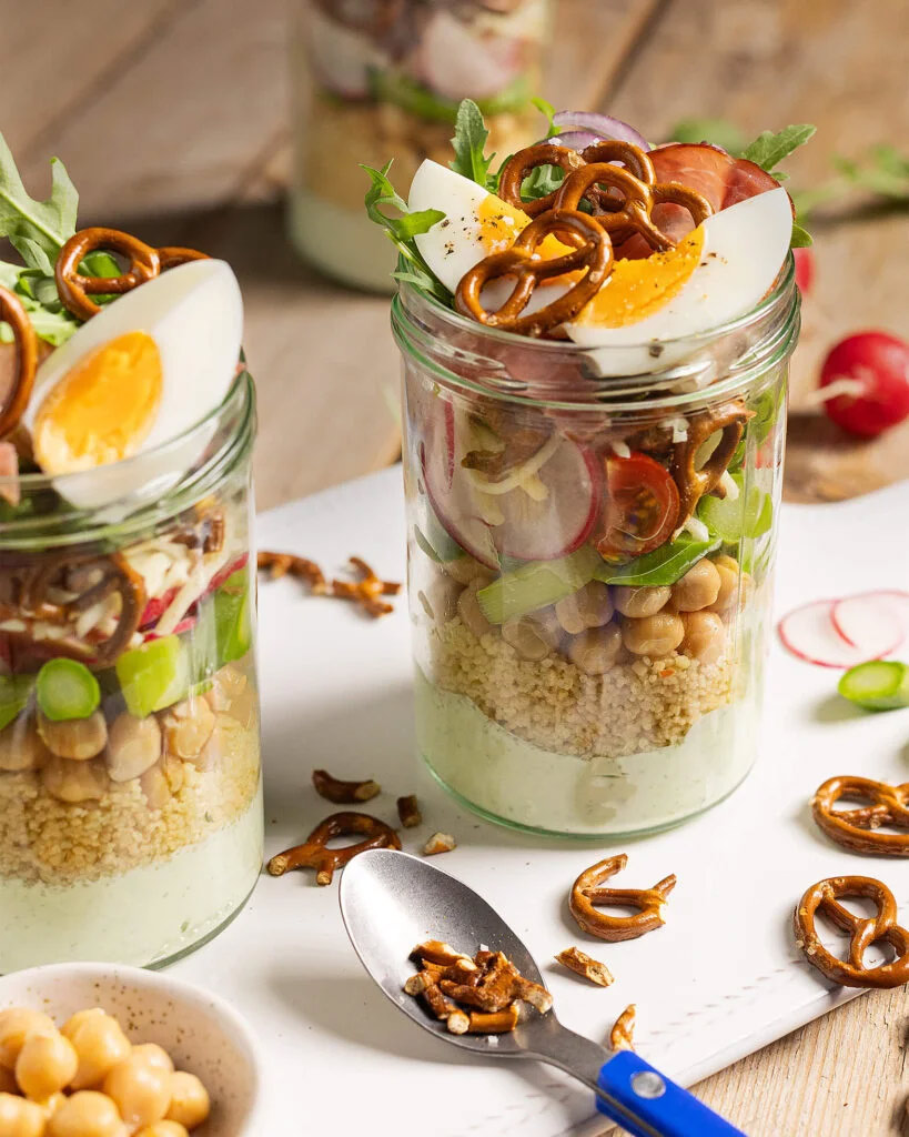 Foto von zwei kleinen Einmachgläsern, gefüllt mit verschiedenen Lagen Salat und mit Eiern und Brezeln als Topping.
