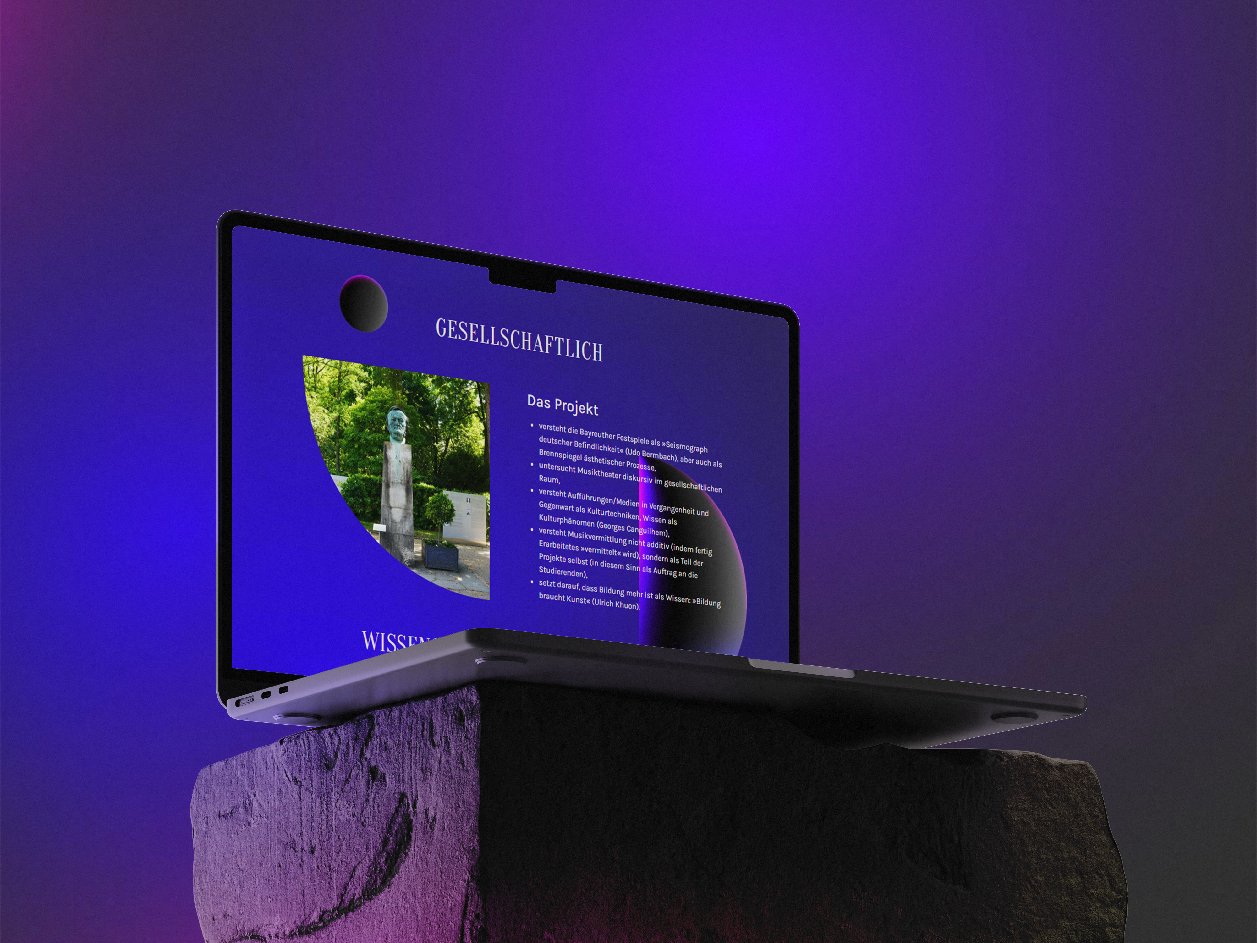 Ein aufgeklappter Laptop steht seitlich auf einem rauen Steinpodest vor einem violett beleuchteten Hintergrund. Auf dem Bildschirm ist eine Website mit der Überschrift „Gesellschaftlich“ zu sehen, inklusive eines Fotos einer Statue und erläuterndem Text zum Projekt.